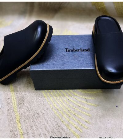 A pair of black leather loafers with tan trim, positioned elegantly on a gray box with the Timberland logo, set against a beige background with yellow accents.