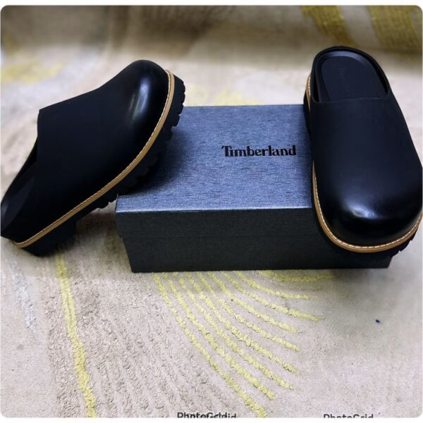 A pair of black leather loafers with tan trim, positioned elegantly on a gray box with the Timberland logo, set against a beige background with yellow accents.