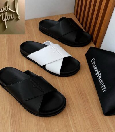A pair of black and white slides on a wooden table next to a black bag with the brand name Cesare Paciotti.