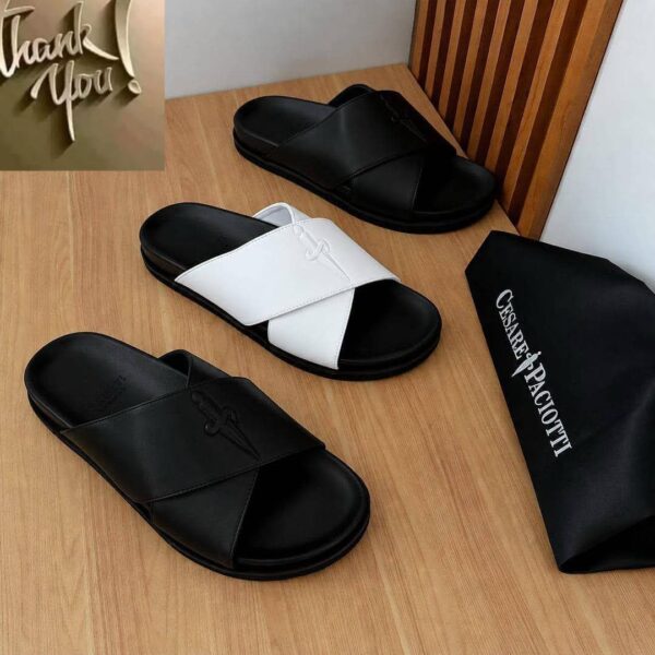 A pair of black and white slides on a wooden table next to a black bag with the brand name Cesare Paciotti.