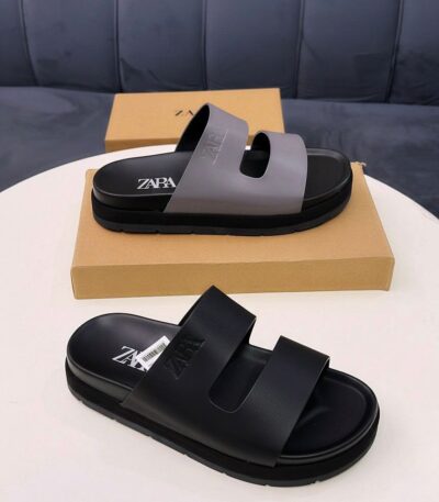 A pair of premium ZARA slides, one in black and the other in black and grey, displayed on a white table with a brown shoe box, exuding luxury and comfort.