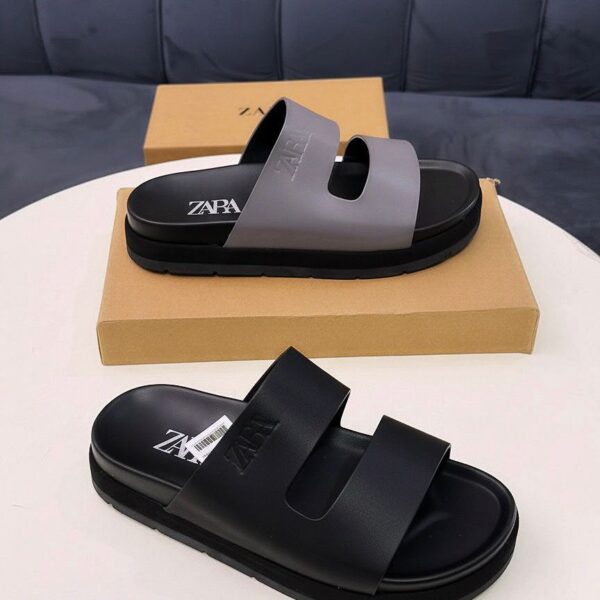 A pair of premium ZARA slides, one in black and the other in black and grey, displayed on a white table with a brown shoe box, exuding luxury and comfort.