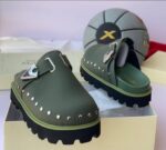 A pair of luxurious green loafers with silver studs and a sleek buckle, displayed on a cream-colored shoe box with a stylish gray and black soccer ball in the background.