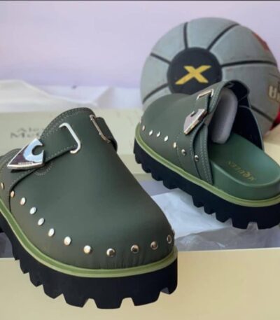 A pair of luxurious green loafers with silver studs and a sleek buckle, displayed on a cream-colored shoe box with a stylish gray and black soccer ball in the background.