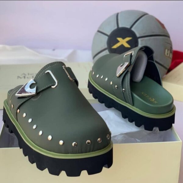 A pair of luxurious green loafers with silver studs and a sleek buckle, displayed on a cream-colored shoe box with a stylish gray and black soccer ball in the background.