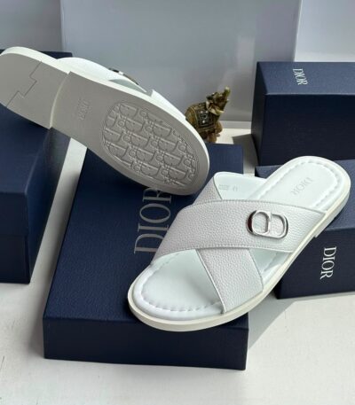 A pair of white leather slides with a silver 'CD' buckle, displayed on navy blue boxes with the 'DIOR' logo.