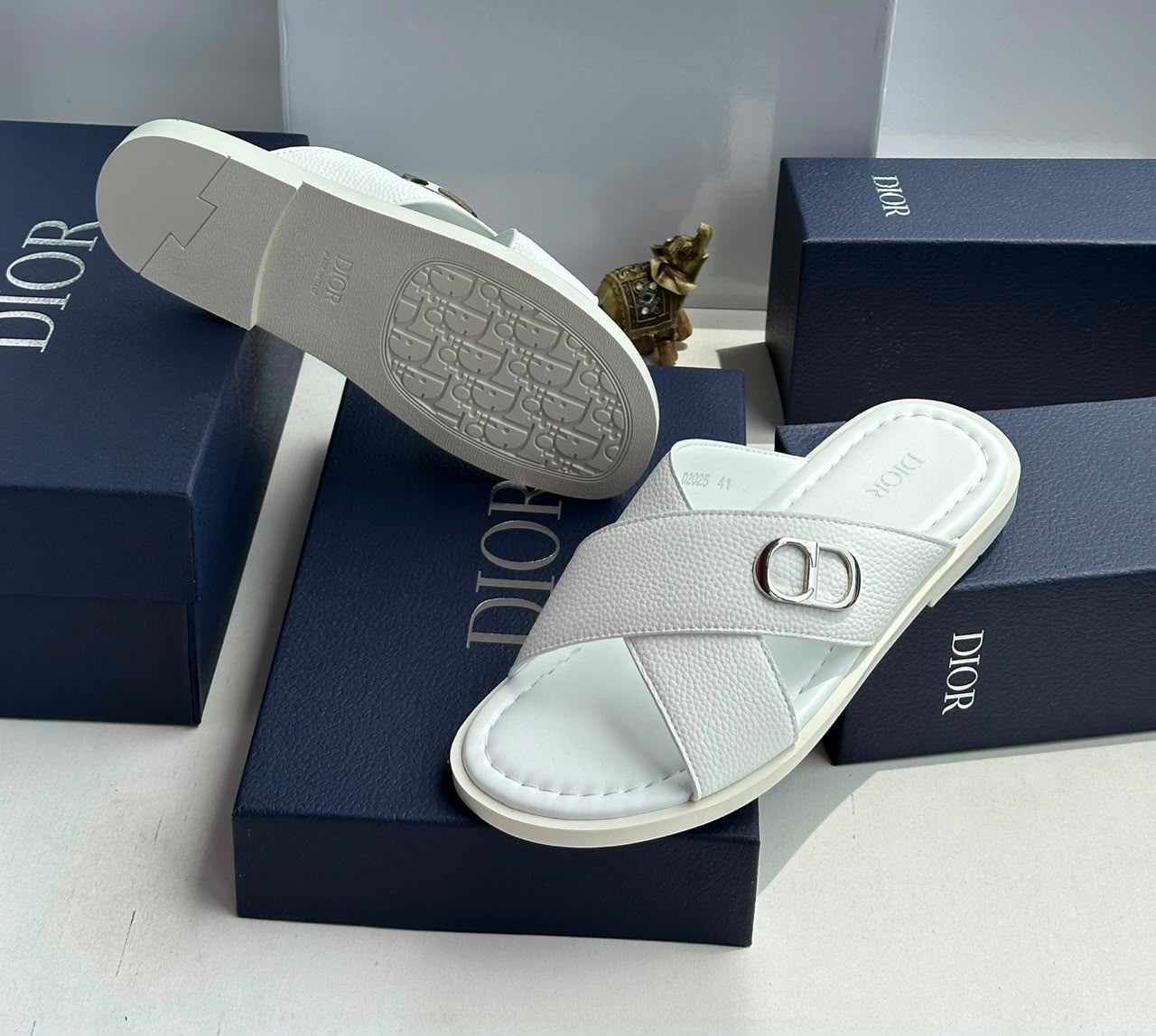 root A pair of white leather slides with a silver 'CD' buckle, displayed on navy blue boxes with the 'DIOR' logo.