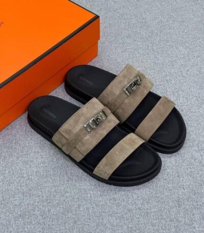 A pair of luxurious black slides with tan suede straps and silver buckles, accompanied by an orange shoe box on a light grey background.