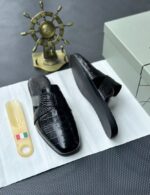 A pair of luxurious black leather half shoes displayed on a white surface, accompanied by a small Italian flag keychain and a stylish gold ship's wheel, evoking a sense of high-end elegance.