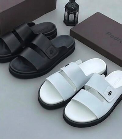 A pair of black and white slides with two wide straps, displayed on a clean surface alongside their brown boxes and a decorative lantern.