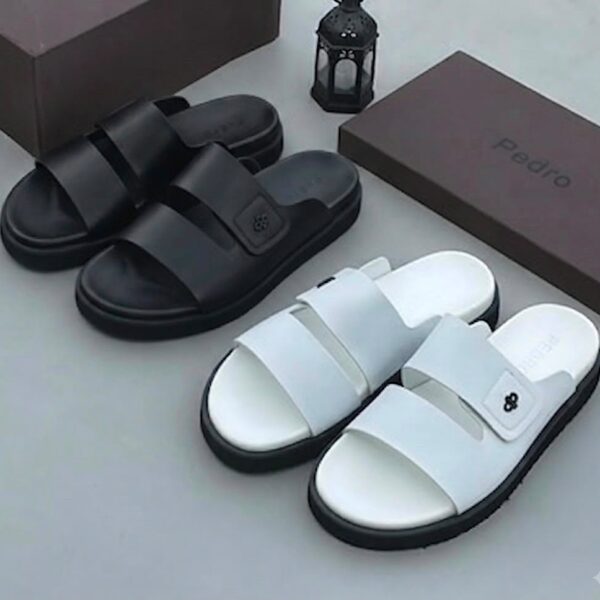 A pair of black and white slides with two wide straps, displayed on a clean surface alongside their brown boxes and a decorative lantern.