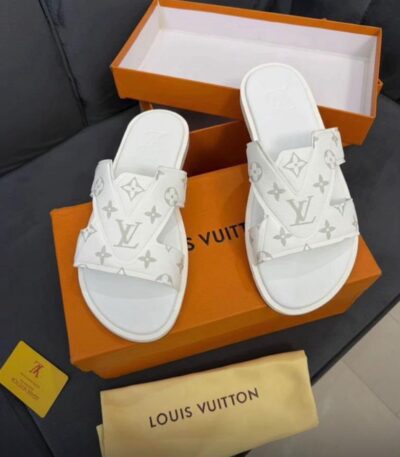 A pair of white Louis Vuitton slides with monogram pattern on a black couch with an open orange box and dust bag.