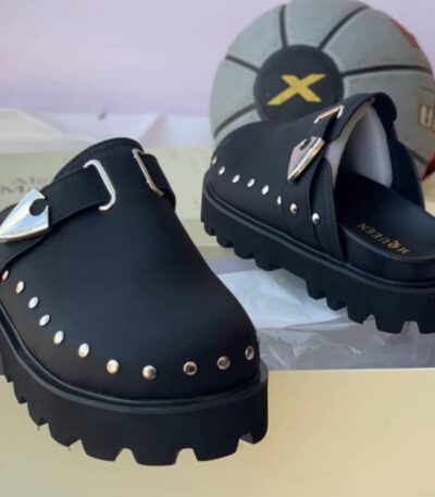 A pair of luxurious green loafers with silver studs and a sleek buckle, displayed on a cream-colored shoe box with a stylish gray and black soccer ball in the background.