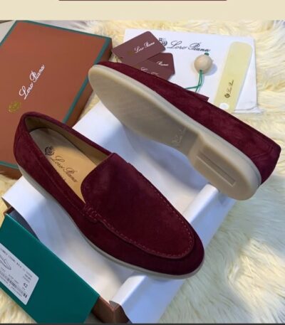 A pair of luxurious, maroon suede loafers showcased on a pristine white box, accompanied by a matching box, dust bag, and shoe care accessories, all set against a plush, cream-colored fur backdrop.